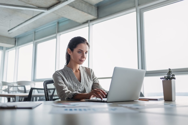 Photo young dark-haired businesswoman wearing striped shirt is sitting in her office and looking into laptop. her notebook and pencil are lying on the desk. bright modern office.