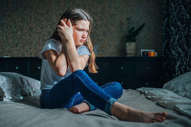 Young dark-haired girl clings to her sore ear with her hands. A girl in a white blouse and blue jeans sits on a bed in her room.