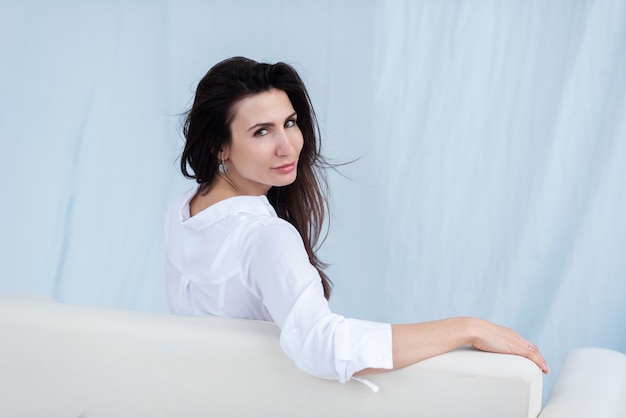 Young dark haired girl in a white shirt and jeans sitting on a white couch back view