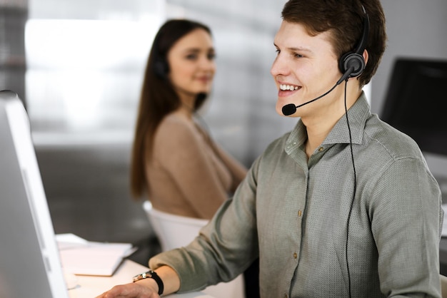 Young dark-haired guy in a green shirt and headsets is talking to a client, while sitting at the desk, working together with a female colleague in a modern office. Call center operators at work.