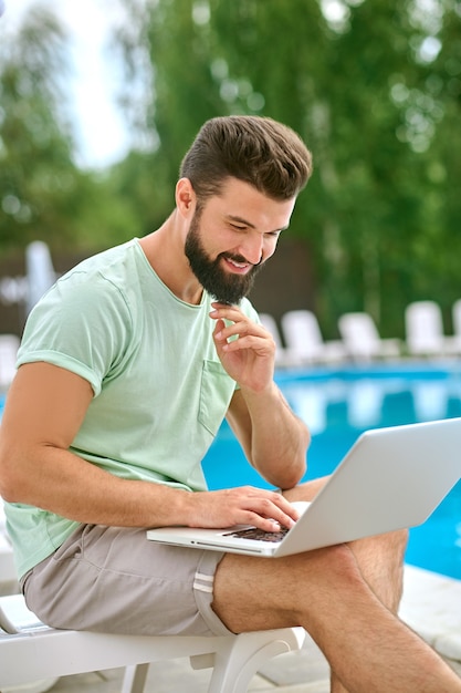 A young dark-haired man working on a laptop while sitting near the swimming pool