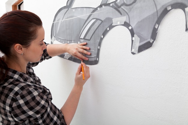 A young dark-haired woman in a plaid shirt draws a large red car in the boy's room on the wall.