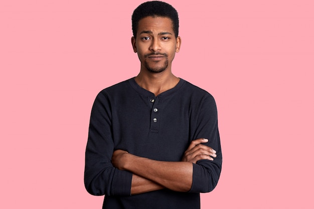 Young dark skinned man with calm expression, short hairstyle and beard, dresses casual shirt and keeps hands crossed. Black male, poses isolated over pink wall. People concept.