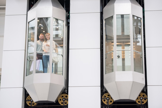 Young dates standing in one of elevators and looking through window while enjoying shopping in the mall on black friday