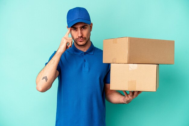 Young delivery caucasian man isolated on blue background pointing temple with finger, thinking, focused on a task.