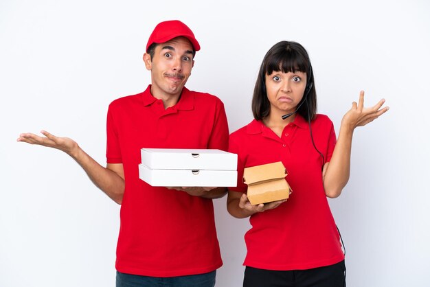 Young delivery couple holding pizzas and burgers isolated on white background having doubts while raising hands and shoulders