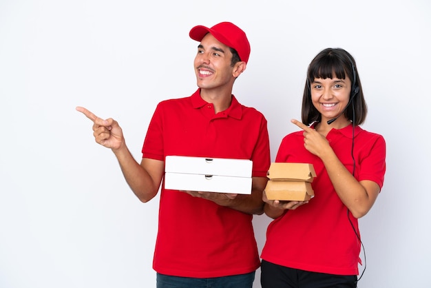 Young delivery couple holding pizzas and burgers isolated on white background pointing finger to the side in lateral position