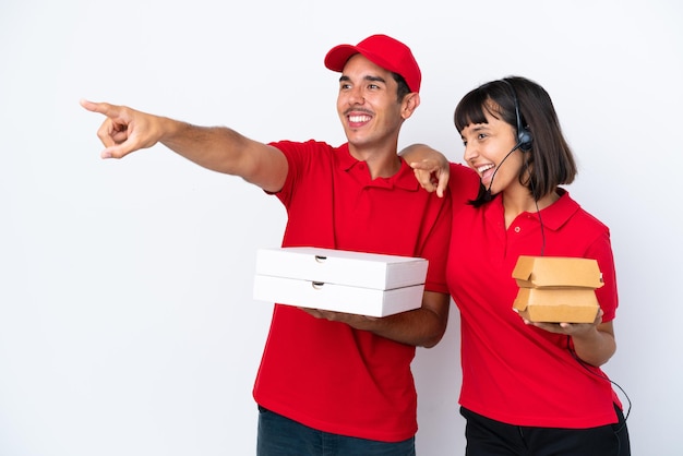 Young delivery couple holding pizzas and burgers isolated on white background pointing to the side to present a product