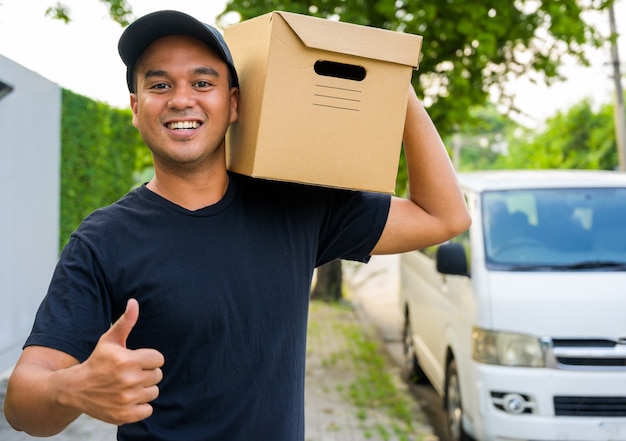 Молодой работник доставляющий покупки на дом в черной униформе выставки thumbup и держать бумажную коробку.