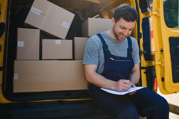 Giovane corriere uomo di consegna in uniforme tenere documenti appunti elenco di controllo pacchi scatole postali vicino a un'auto per il servizio di spedizione al cliente concetti di servizio di acquisto online