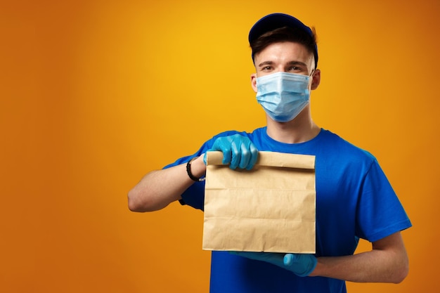 Young delivery man in face mask and gloves holding food delivery parcel against yellow background