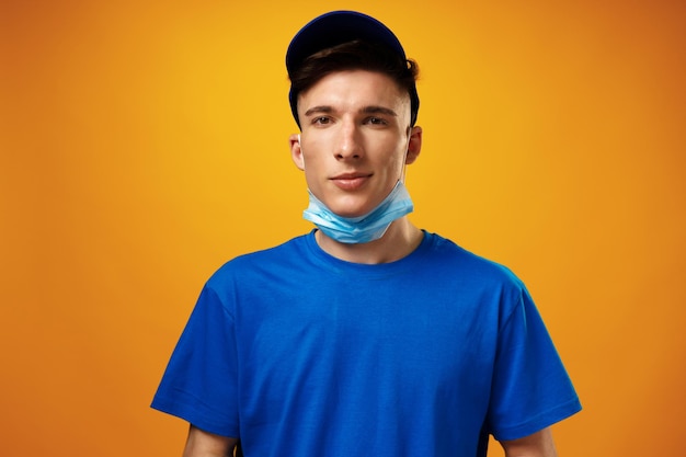 Young delivery man in green tshirt and cap wearing face mask against yellow background