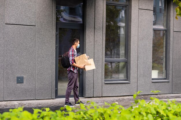 Photo young delivery man in medical mask on face holding paper bags in hands. hipster man courier with shopping bags. safe online delivery service. coronavirus covid-19 protection