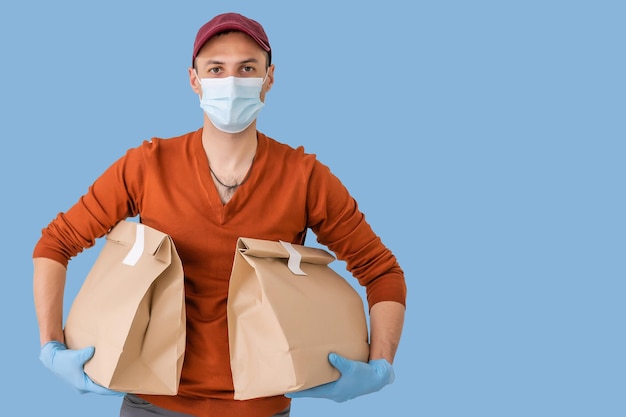 Young delivery man in medical mask holding and carrying a cardbox isolated on blue background. Buy food online in quarantine concept.