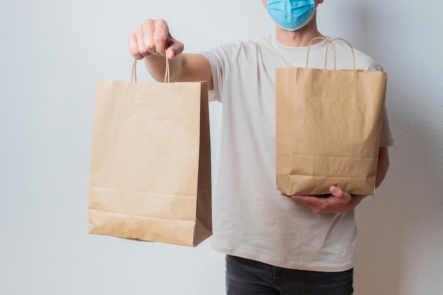 Young delivery man in protective face mask holding paper bags in hands. Man courier with shopping bag. Safe online delivery from supermarket to home. COVID-19 coronavirus protection