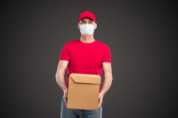 Молодой курьер в красной кепке и футболке с защитной маской держит картонную упаковку с заказанным продуктом во время работы во время вспышки коронавируса