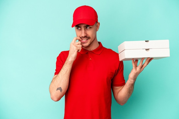 Young delivery man taking pizzas isolated on blue bakcground relaxed thinking about something looking at a copy space.