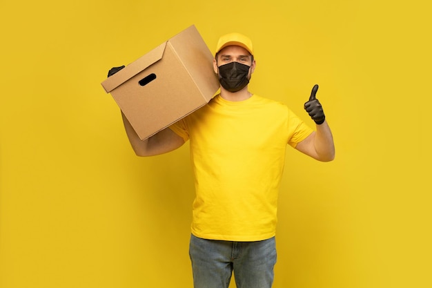 Young delivery man with box in mask isolated over yellow background coronavirus 2019 concept