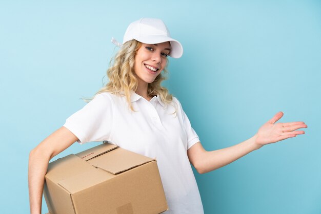 Young delivery woman on blue extending hands to the side for inviting to come