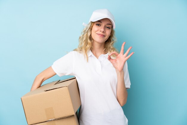 Young delivery woman on blue showing an ok sign with fingers