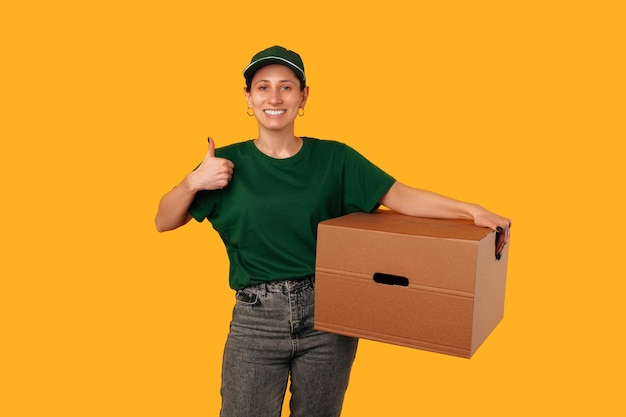 Young delivery woman is holding a big moving box while showing thumb up