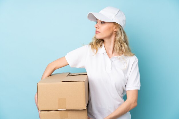 Young delivery woman isolated on blue wall looking side