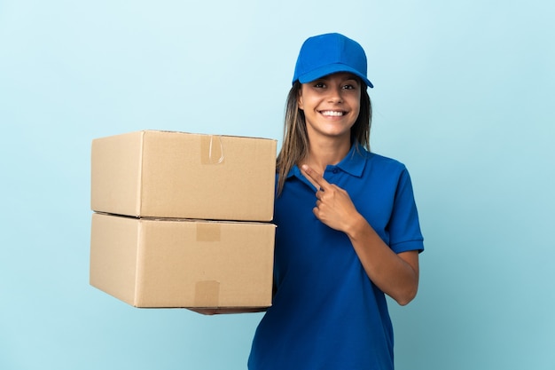 Young delivery woman isolated on blue wall pointing to the side to present a product