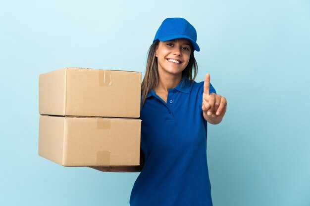 Young delivery woman isolated on blue wall showing and lifting a finger