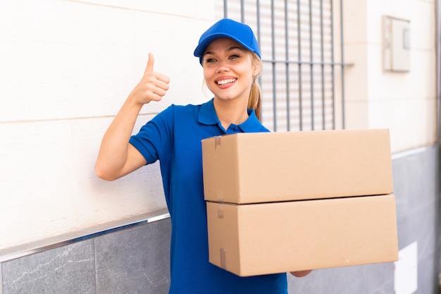 Young delivery woman at outdoors holding boxes with thumb up