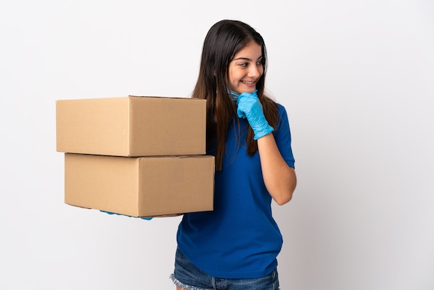 Young delivery woman protecting from the coronavirus with a mask isolated on white wall thinking an idea and looking side