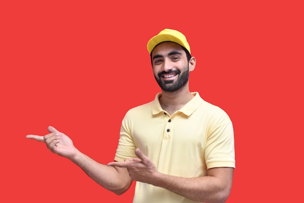 young delivery in yellow tshirt smiling pointing aside over red background indain pakistani model