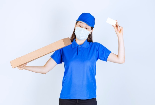 Young deliverywoman in medical mask holding package and business card. 