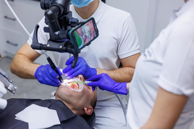 Foto un giovane dentista esamina i denti del paziente con un microscopio dentale e tiene gli strumenti dentali vicino alla bocca un assistente aiuta il medico indossano uniformi bianche con maschere