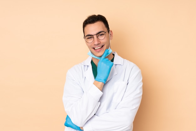 Young dentist man holding tools on beige wall with glasses and happy