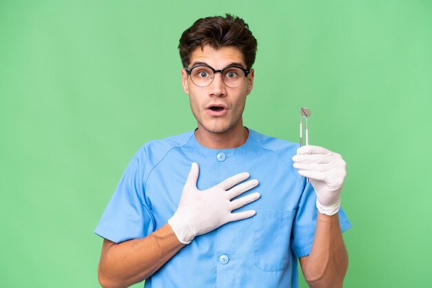 Young dentist man holding tools over isolated background surprised and shocked while looking right