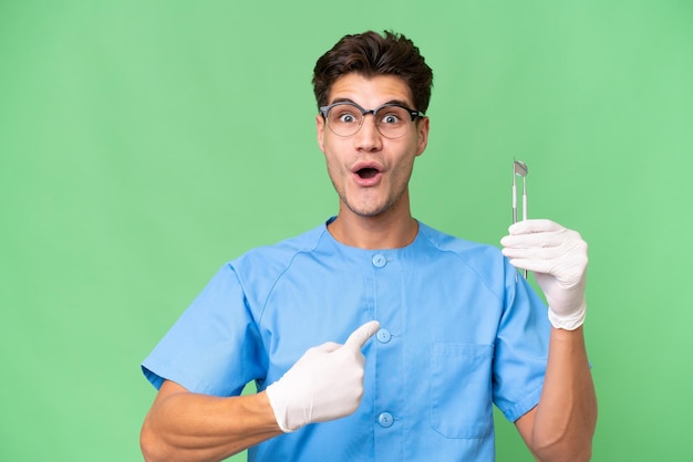 Young dentist man holding tools over isolated background with surprise facial expression