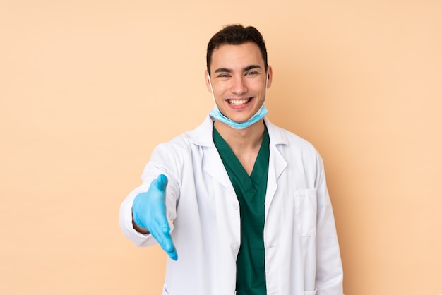 Молодой стоматолог мужчина держит инструменты, изолированные на бежевой стене рукопожатие для закрытия хорошей сделки