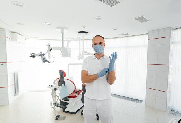 A young dentist in a mask stands near a red dental chair and smiles in modern white dentistry Modern dentistry and prosthetics