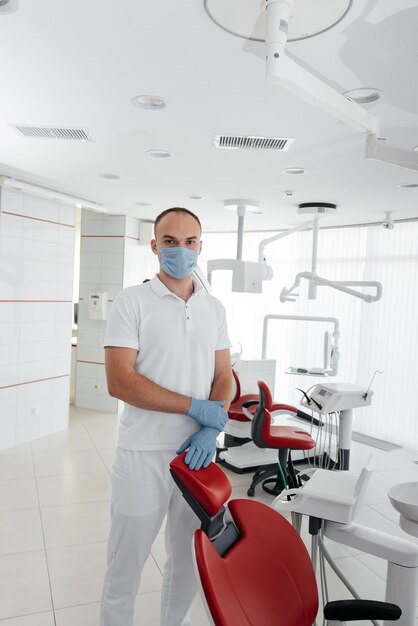 A young dentist in a mask stands near a red dental chair and smiles in modern white dentistry Modern dentistry and prosthetics