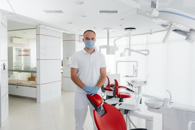 A young dentist in a mask stands near a red dental chair and smiles in modern white dentistry Modern dentistry and prosthetics