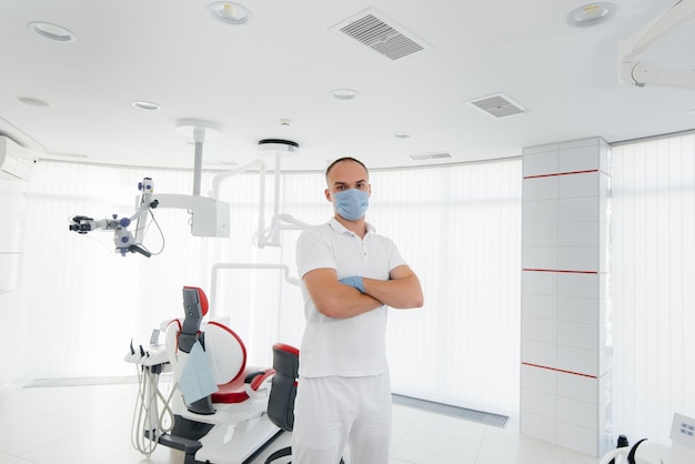 A young dentist in a mask stands near a red dental chair and smiles in modern white dentistry Modern dentistry and prosthetics