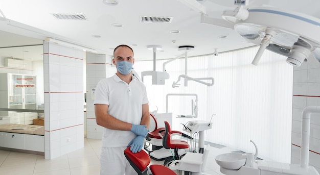 A young dentist in a mask stands near a red dental chair and\
smiles in modern white dentistry modern dentistry and\
prosthetics