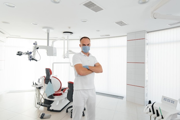 A young dentist in a mask stands near a red dental chair and smiles in modern white dentistry Modern dentistry and prosthetics