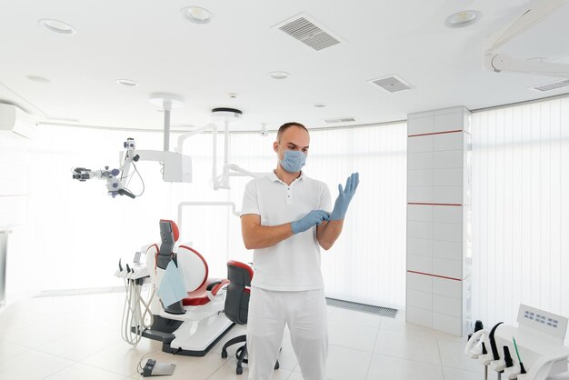 A young dentist in a mask stands near a red dental chair and smiles in modern white dentistry Modern dentistry and prosthetics