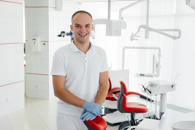 A young dentist stands near a red dental chair and smiles in modern white dentistry Treatment and prevention of caries from youth Modern dentistry and prosthetics