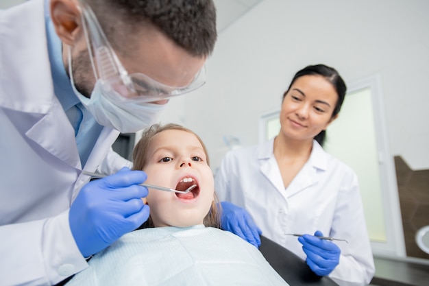 Young dentist using dental mirror during oral examination of little girl keeping her mouth open