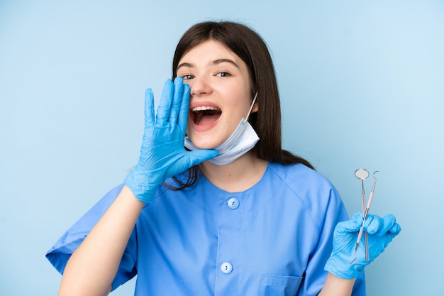 Giovani strumenti di tenuta della donna del dentista sopra la parete blu isolata che grida con la bocca spalancata