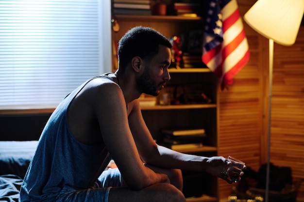 Young depressed man with glass of whisky sitting on bed in bedroom