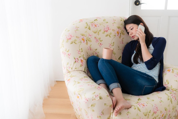 窓のソファの椅子の前に座って泣いている若い落ち込んだ女性は、携帯電話のスマートフォンを使って友達を呼んで、苦しんだときに不平を言った。