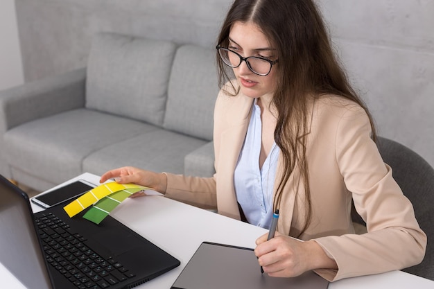 Молодая девушка-дизайнер, сидящая за столом, рисует на планшете, делает бизнес-расчеты на компьютере.